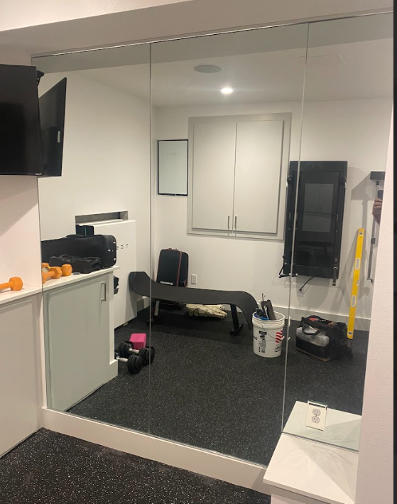Home gym with mirrors on one wall, weights, bench, and exercise equipment. Dark flooring and white walls.