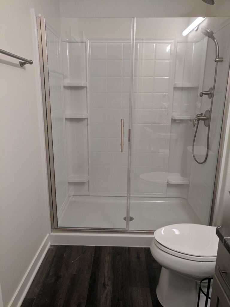 Bathroom with a glass-enclosed shower, toilet, and dark wood-look flooring. White tile-like walls and a silver towel bar.