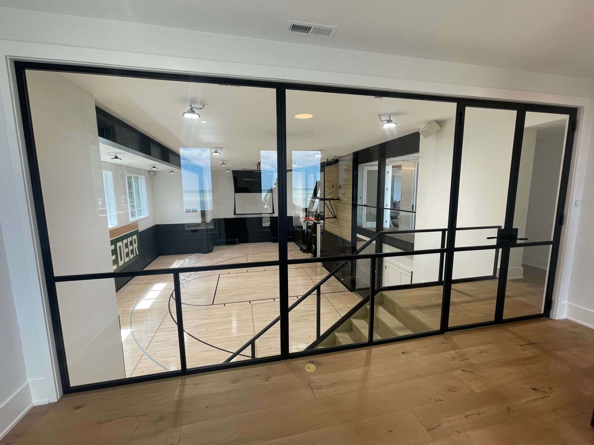 Interior view through black-framed glass wall to a modern kitchen with dark cabinets, light floors.