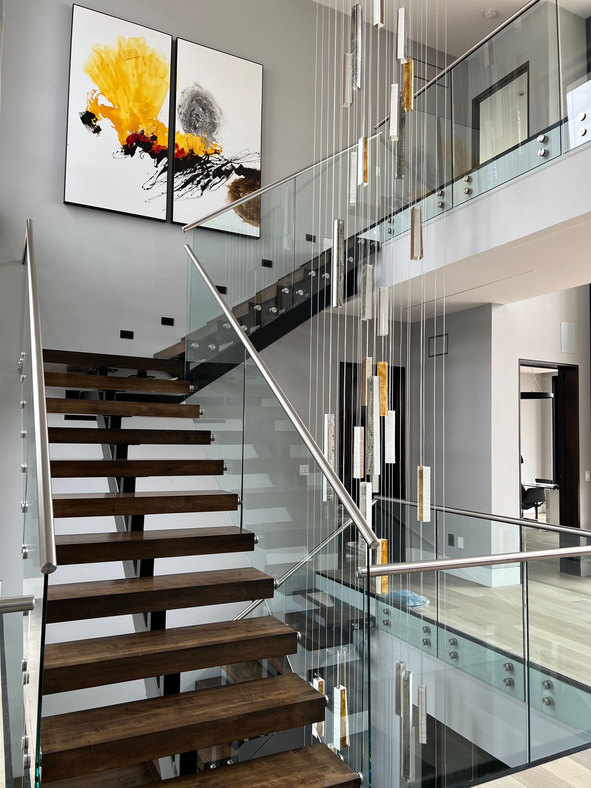 Modern staircase with wooden steps, glass railings, and a chandelier. Abstract art hangs on the wall.