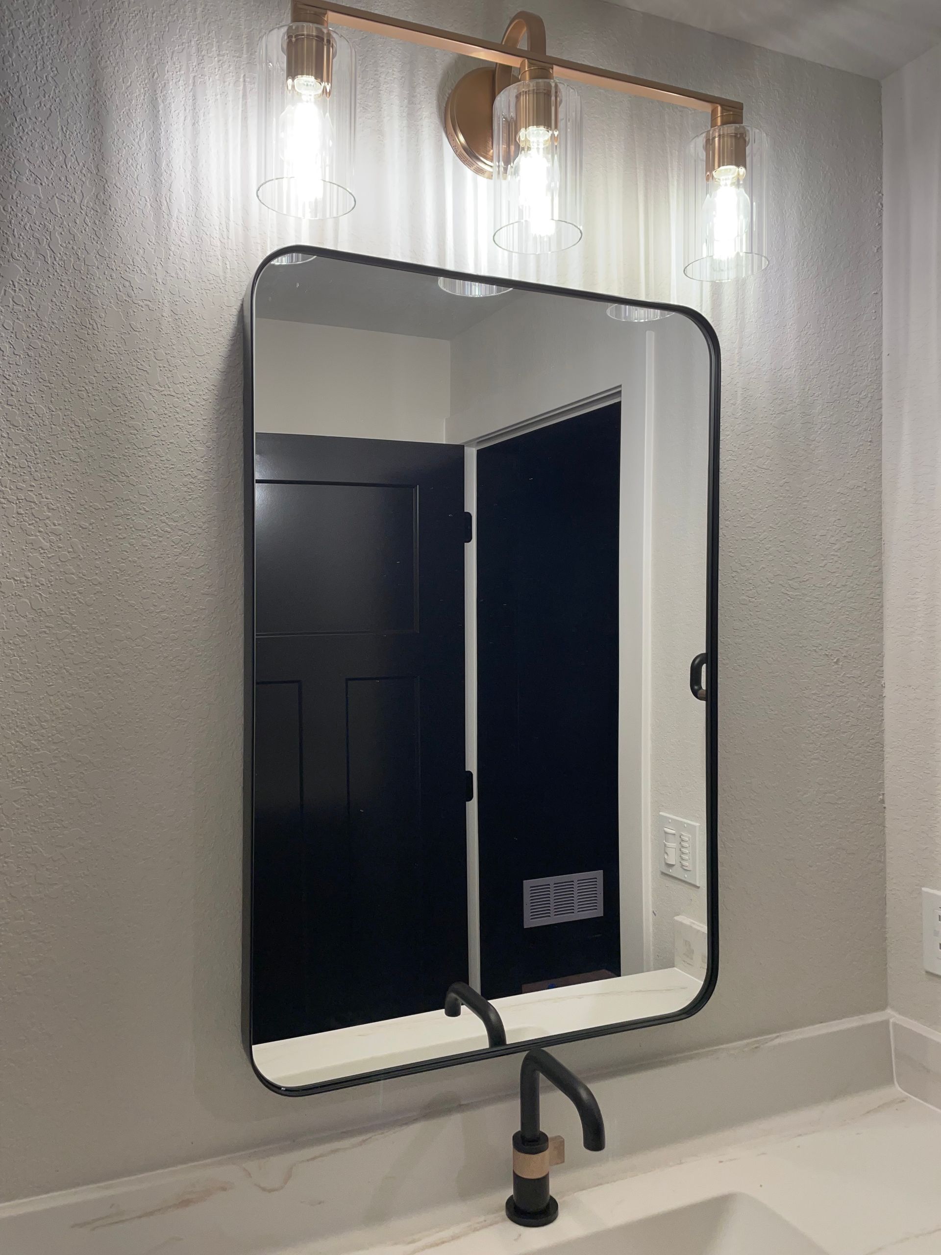 Bathroom vanity with a black-framed mirror, bronze light fixture, black faucet, and a black door in the reflection.