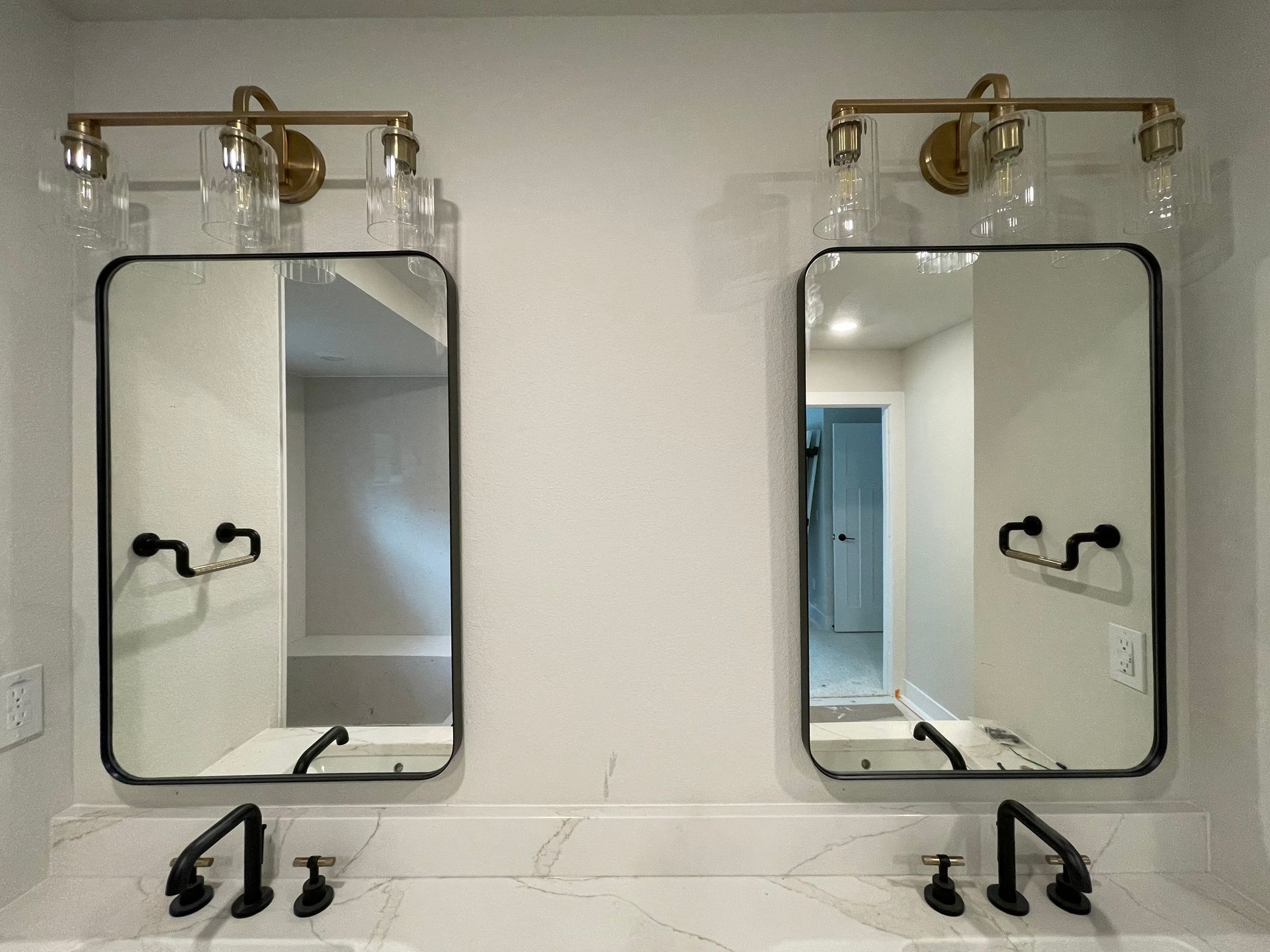 Two vanity mirrors with black frames, sconces, and fixtures on a marble countertop.