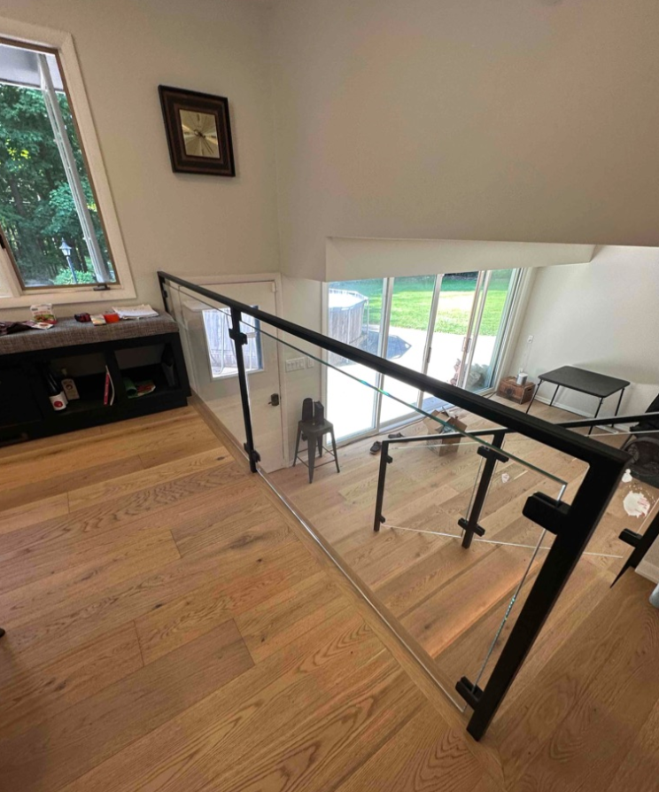 Wooden staircase with glass and black metal railing, leading down to a bright room with a glass door.