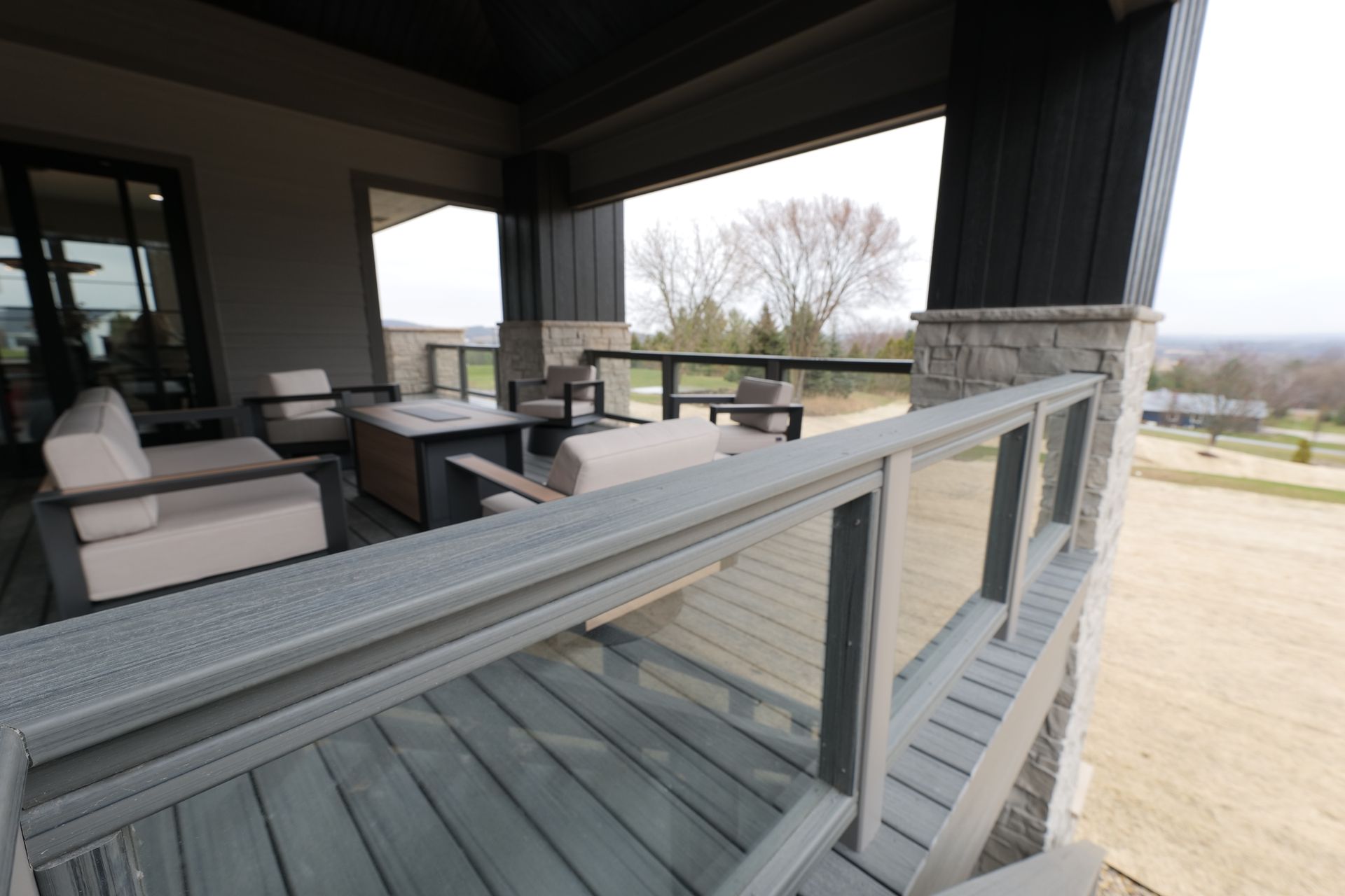 A covered outdoor patio with seating. Glass railing with gray decking, and views of trees and landscape.