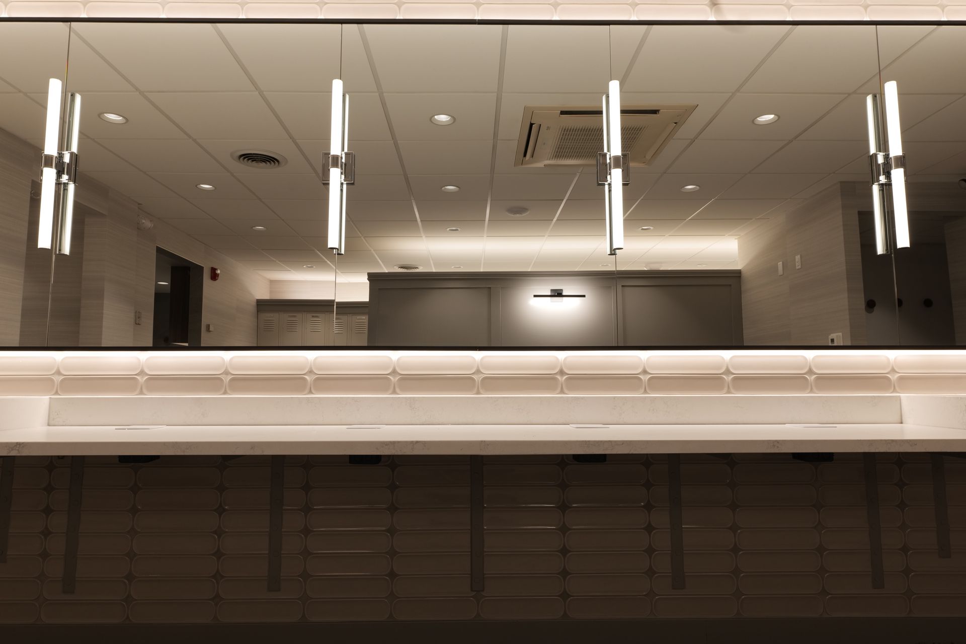 Modern, well-lit interior. A long, light-colored counter with drawers below. Rectangular hanging lights and a mirrored wall.