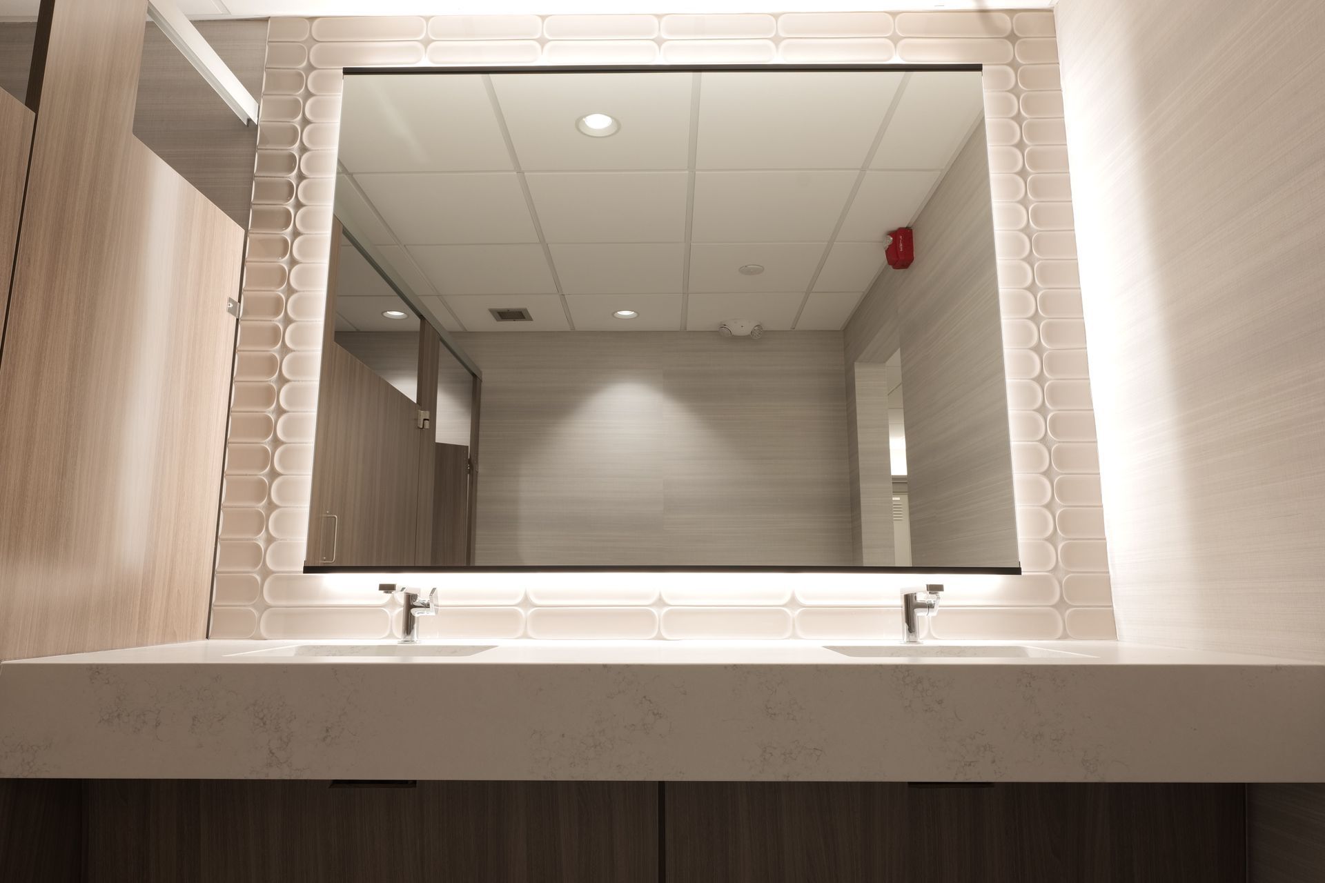 Mirror above a double sink in a public restroom; white countertop, tan wood cabinets, and white tiled wall.