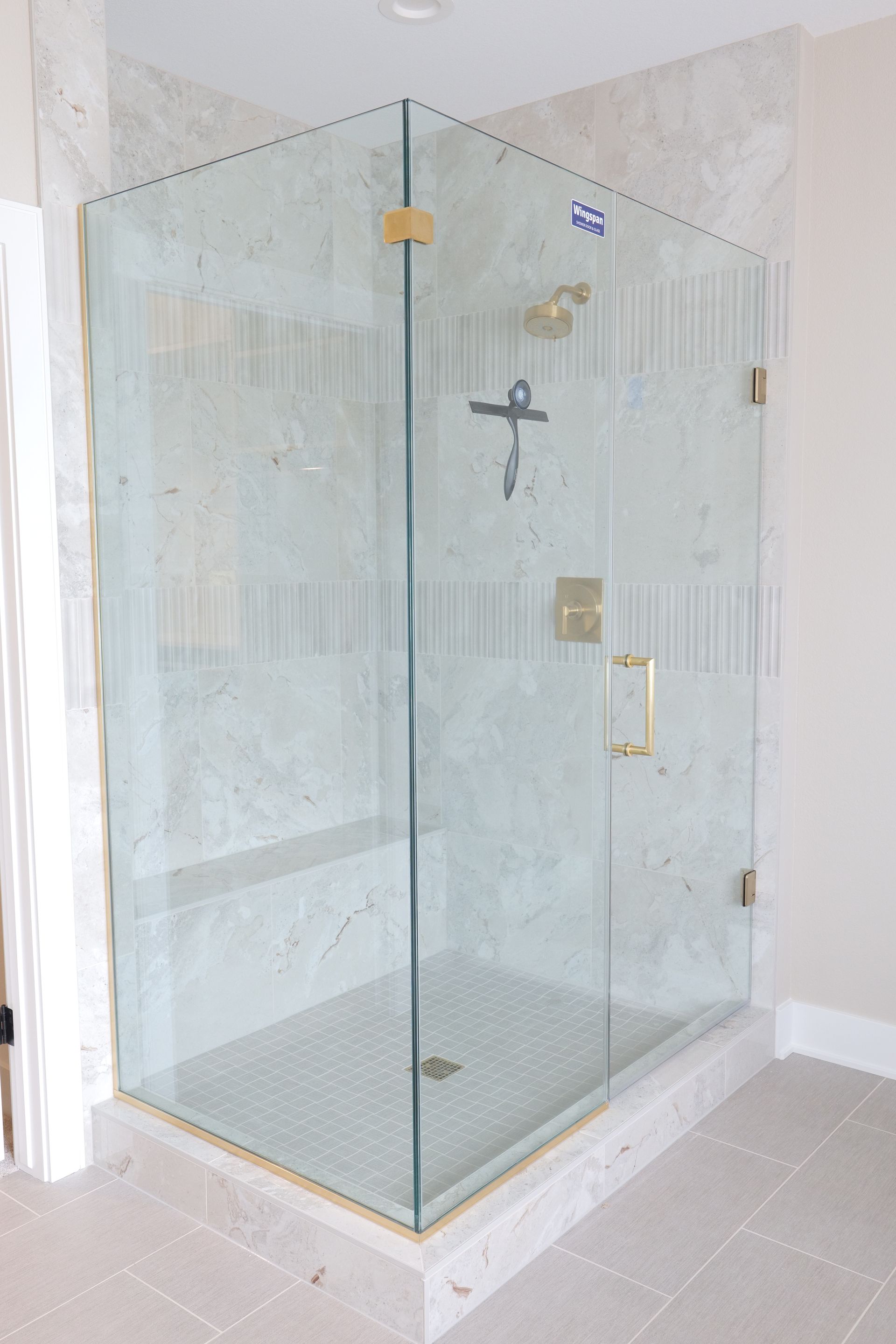 A corner shower enclosure with clear glass doors and gold hardware. The walls are covered in light-colored tiles.