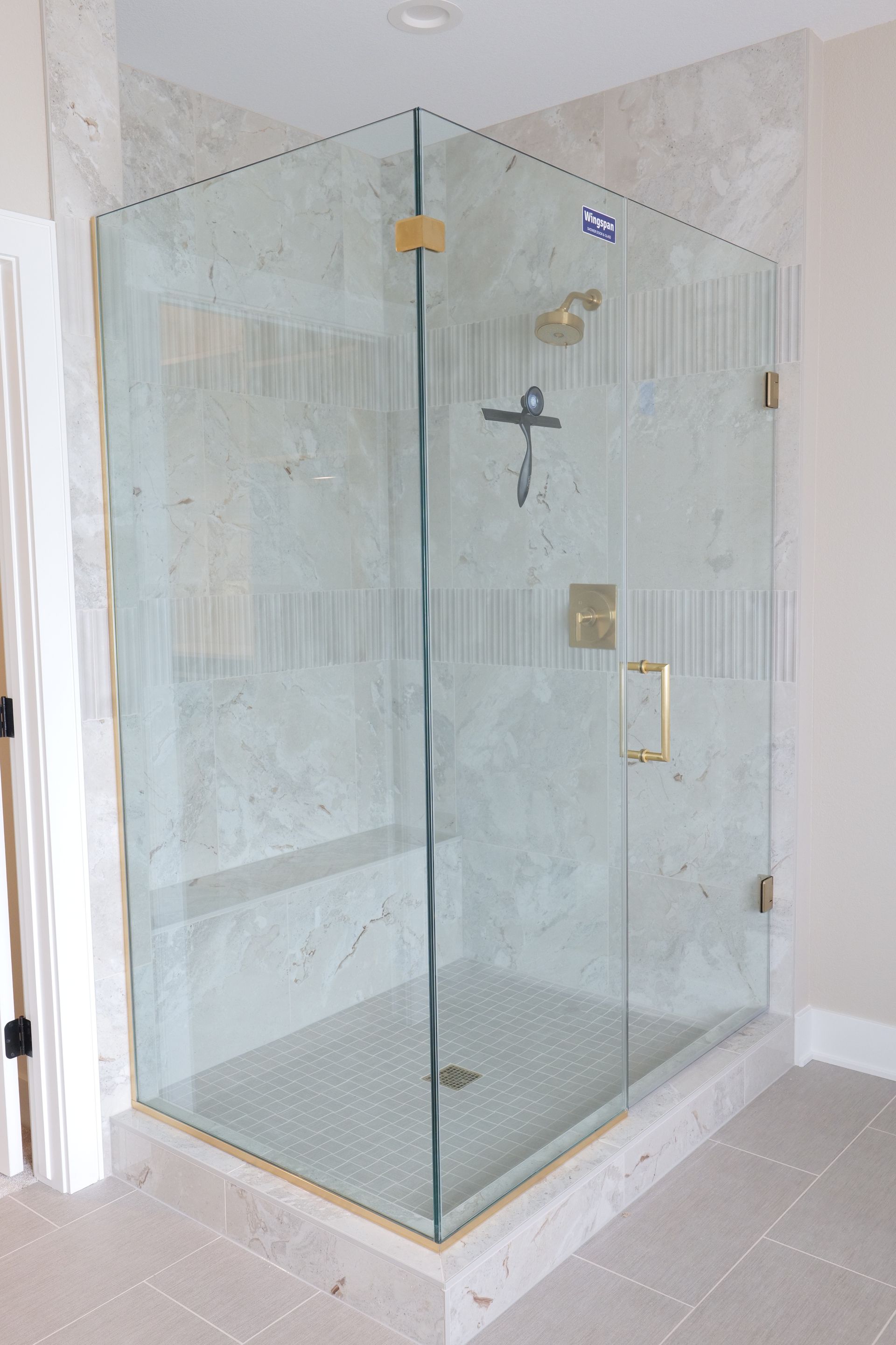 Glass-enclosed corner shower with gold hardware and light-colored tile.