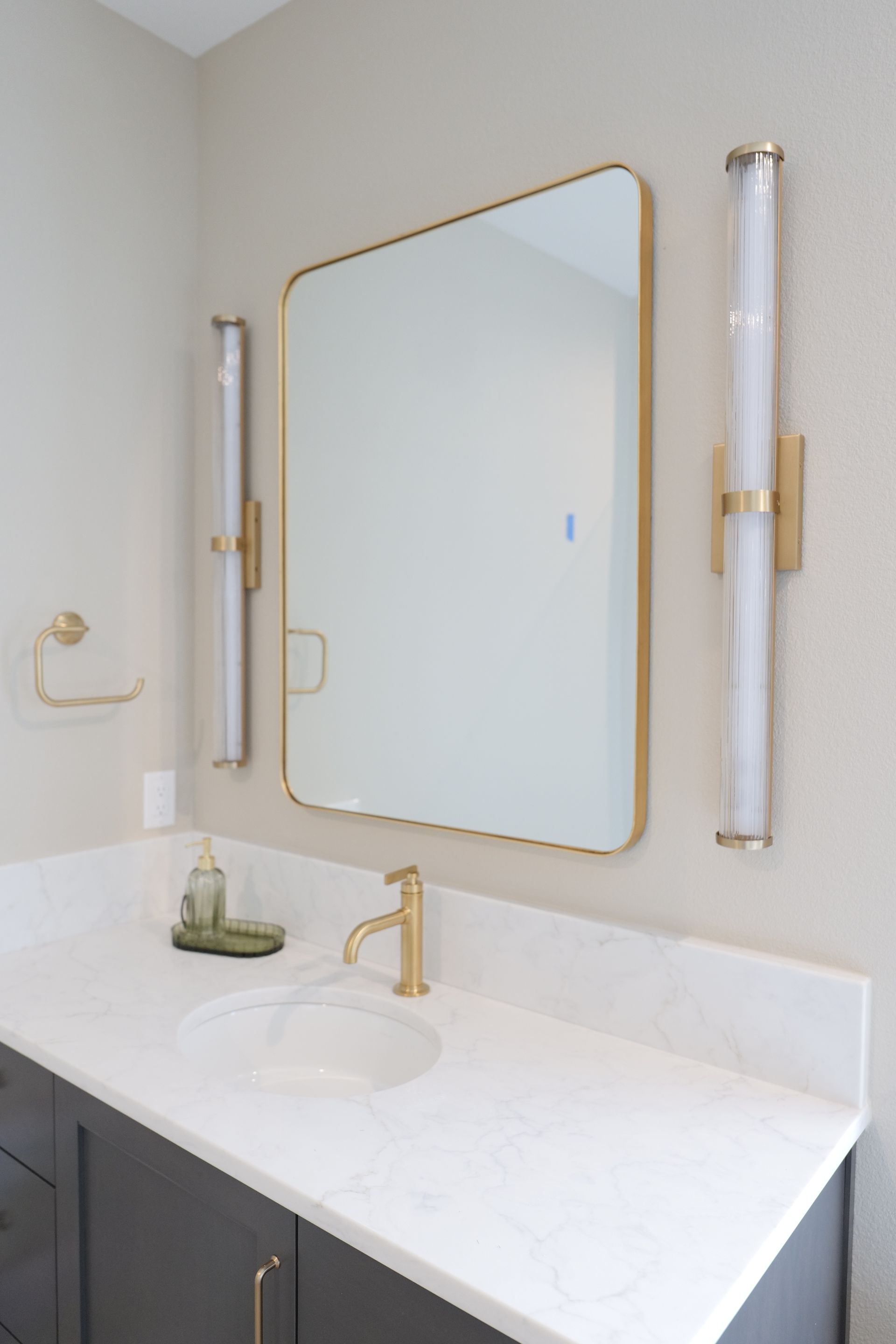Bathroom with gold fixtures, a large mirror, and vanity. White countertop, gray cabinets, and sconces.