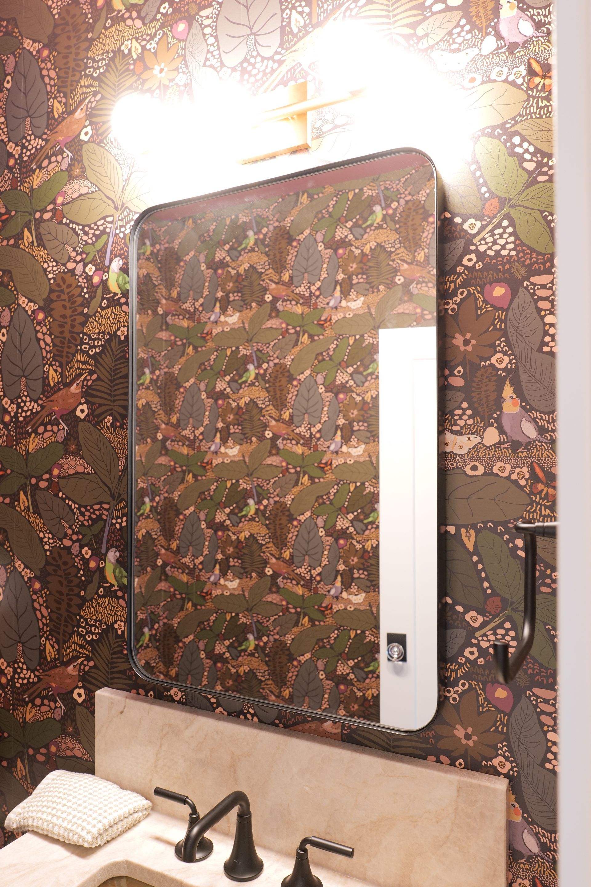 Bathroom mirror with patterned wallpaper background and bronze fixtures.