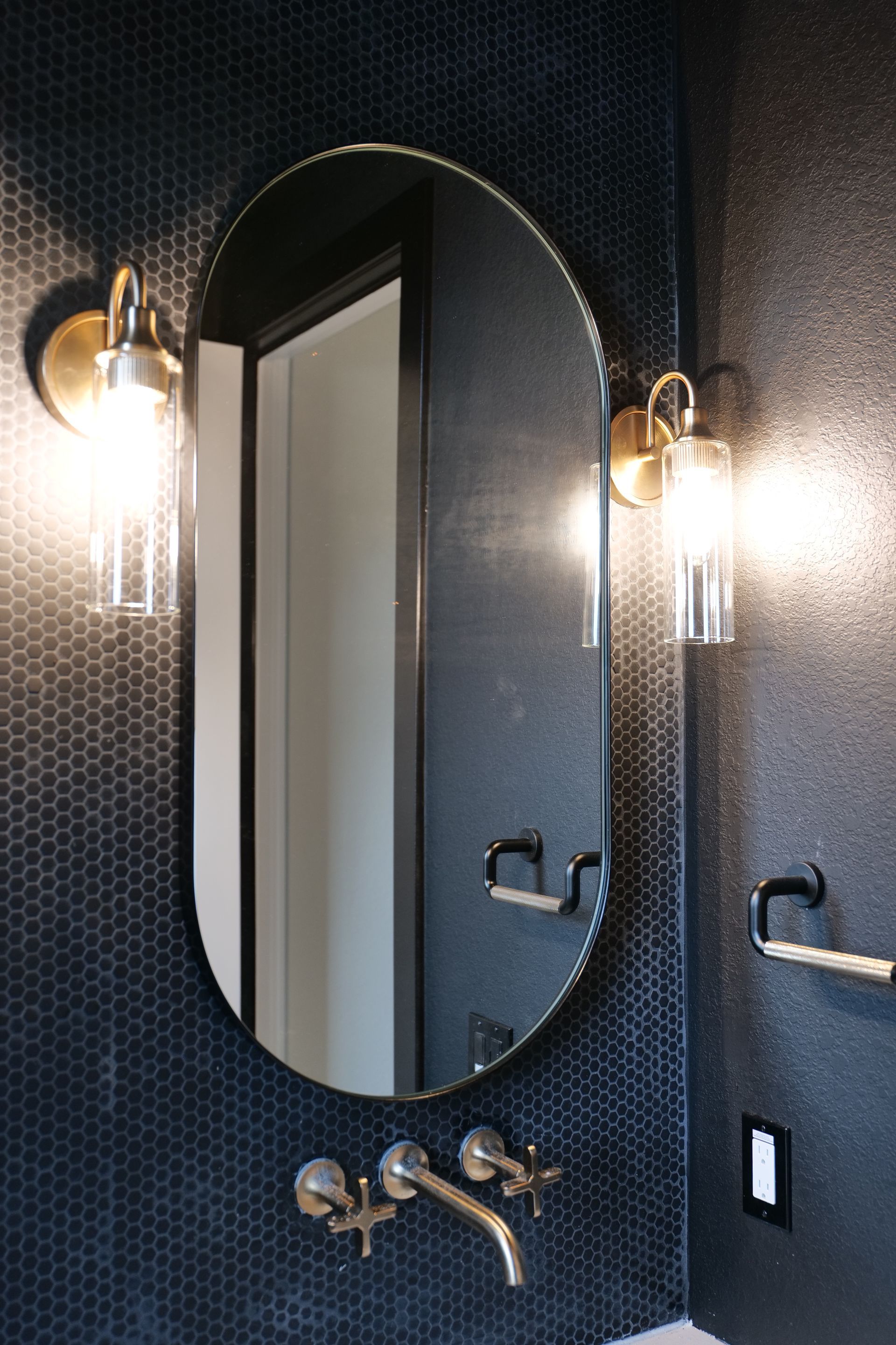 Oval mirror with gold sconces and faucet in a dark blue tiled bathroom.