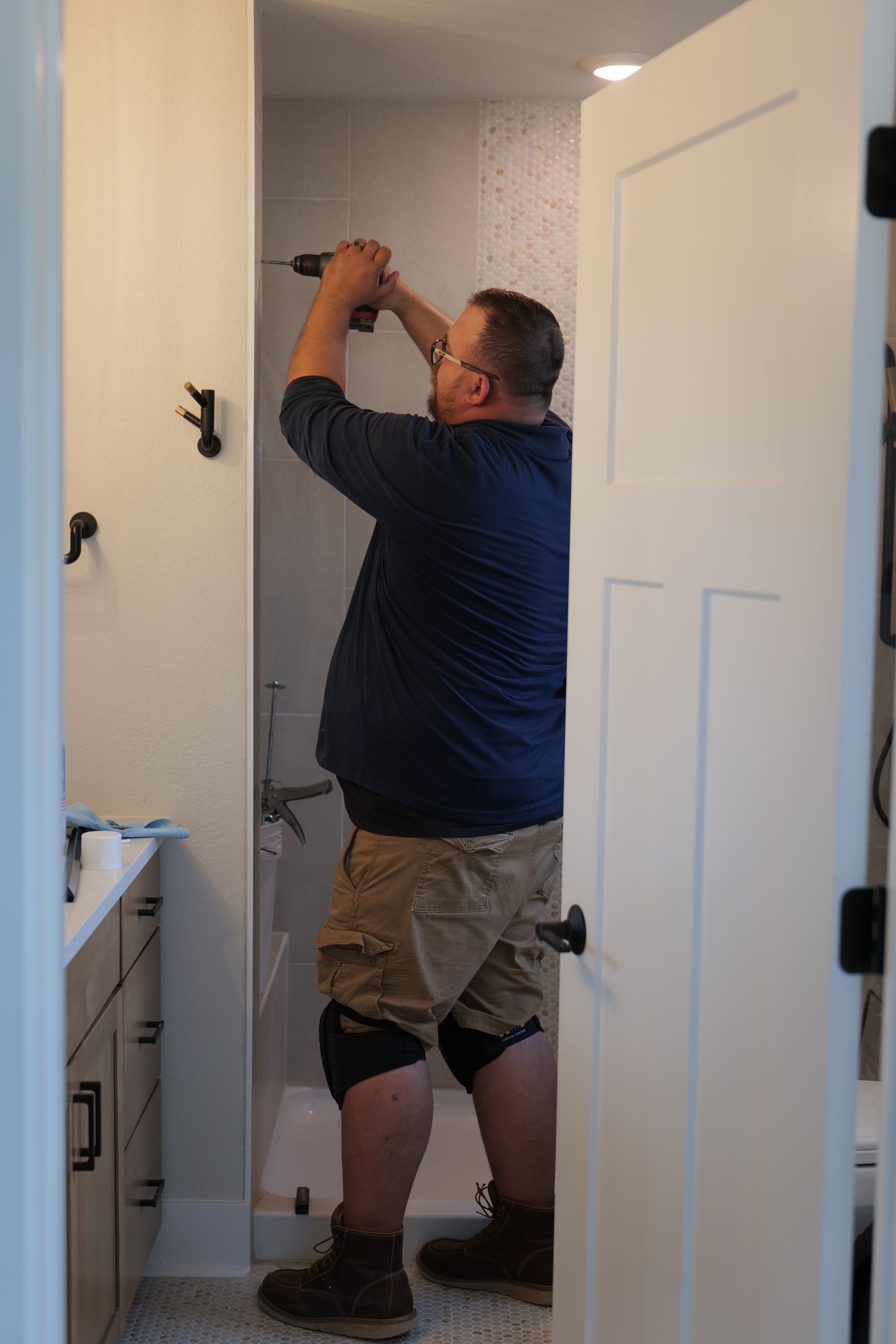 Man installing something with a drill inside a bathroom doorway. He is wearing knee pads, brown work boots, and khaki shorts.