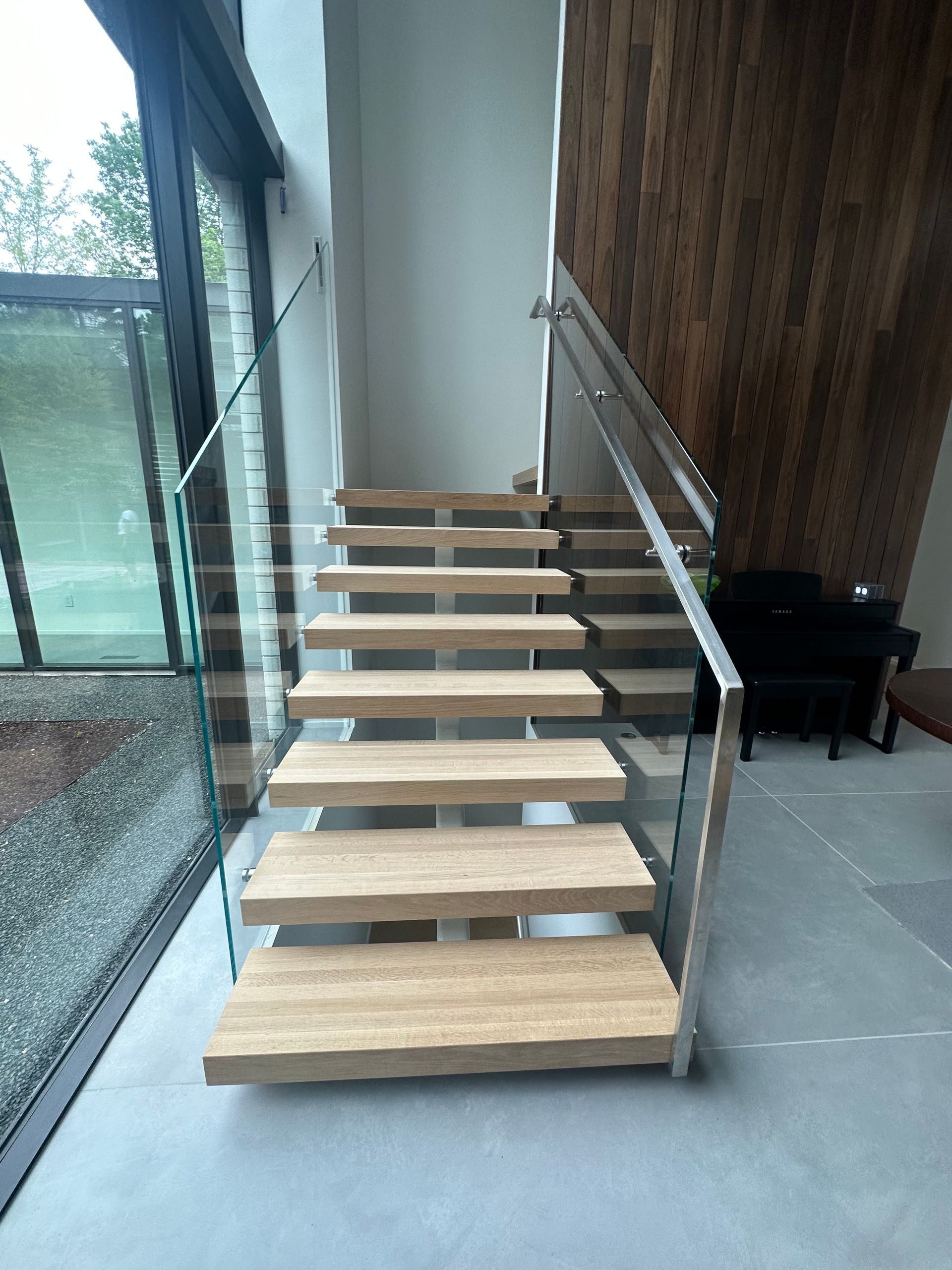Modern wooden staircase with glass railing. Light wood steps, grey concrete floor, and large window.