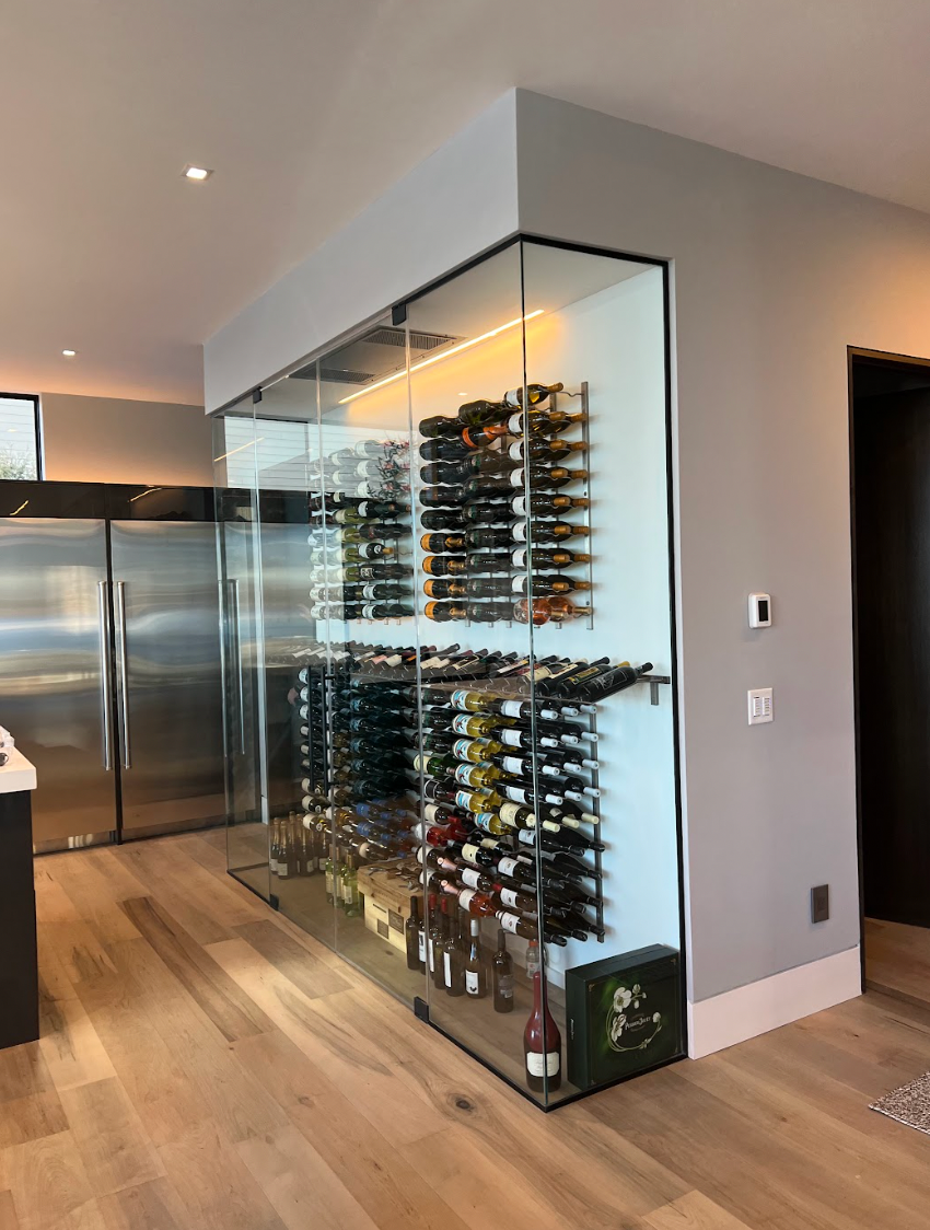 Glass-enclosed wine cellar with stainless steel racks, holding numerous wine bottles, 