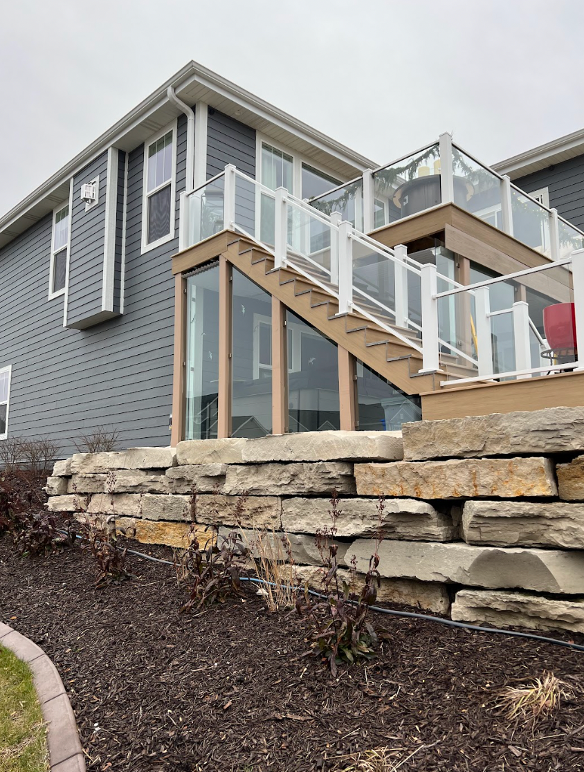 Exterior view of a house with a multi-level deck, glass railings, and a stone retaining wall.