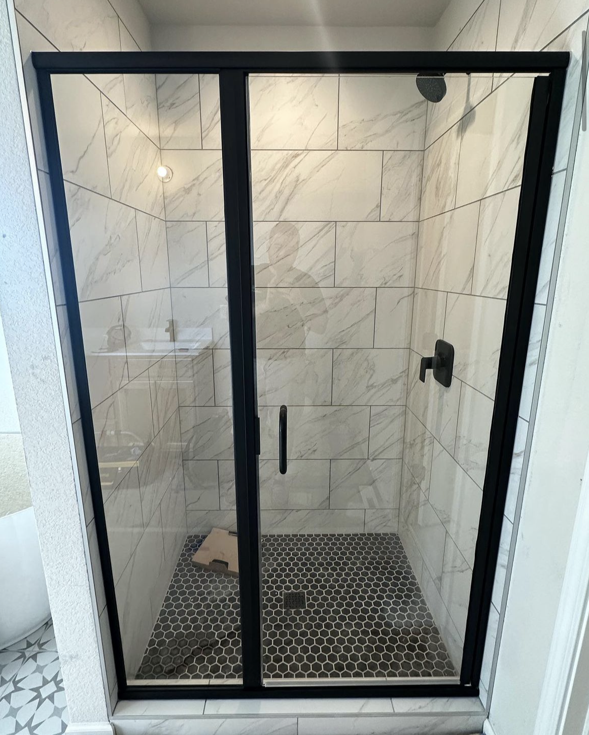 Black-framed shower enclosure with a clear glass door. White marble-look tile walls, dark mosaic floor.