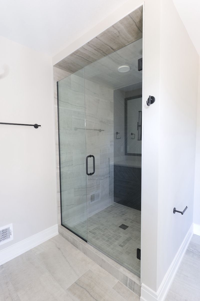Modern shower with glass door, black hardware, gray tile, and light-colored walls.