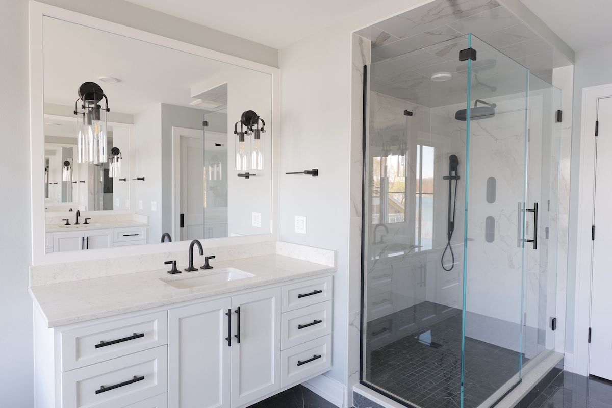 Modern white bathroom with vanity, large mirror, glass shower, and black accents.