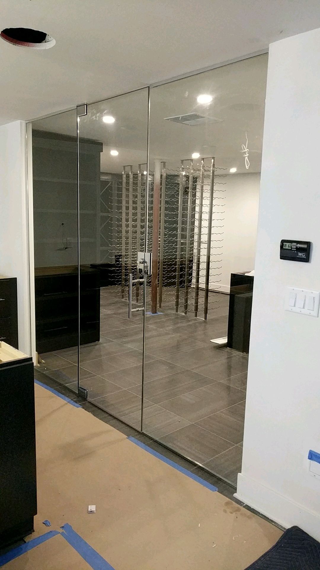 Glass-walled wine cellar with stainless steel racks. Interior view with tiled floor, modern lighting, and open door.