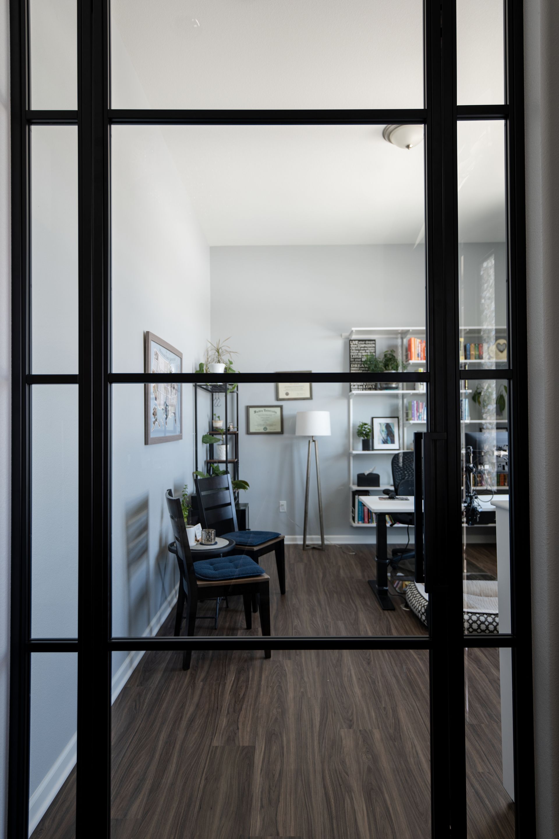 Interior view of a modern home office through black-framed glass doors; two chairs, desk, and shelves are visible.