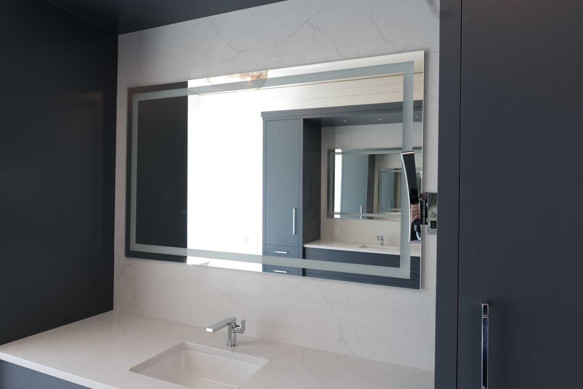 Bathroom with large illuminated mirror over white countertop, sink, and cabinet.