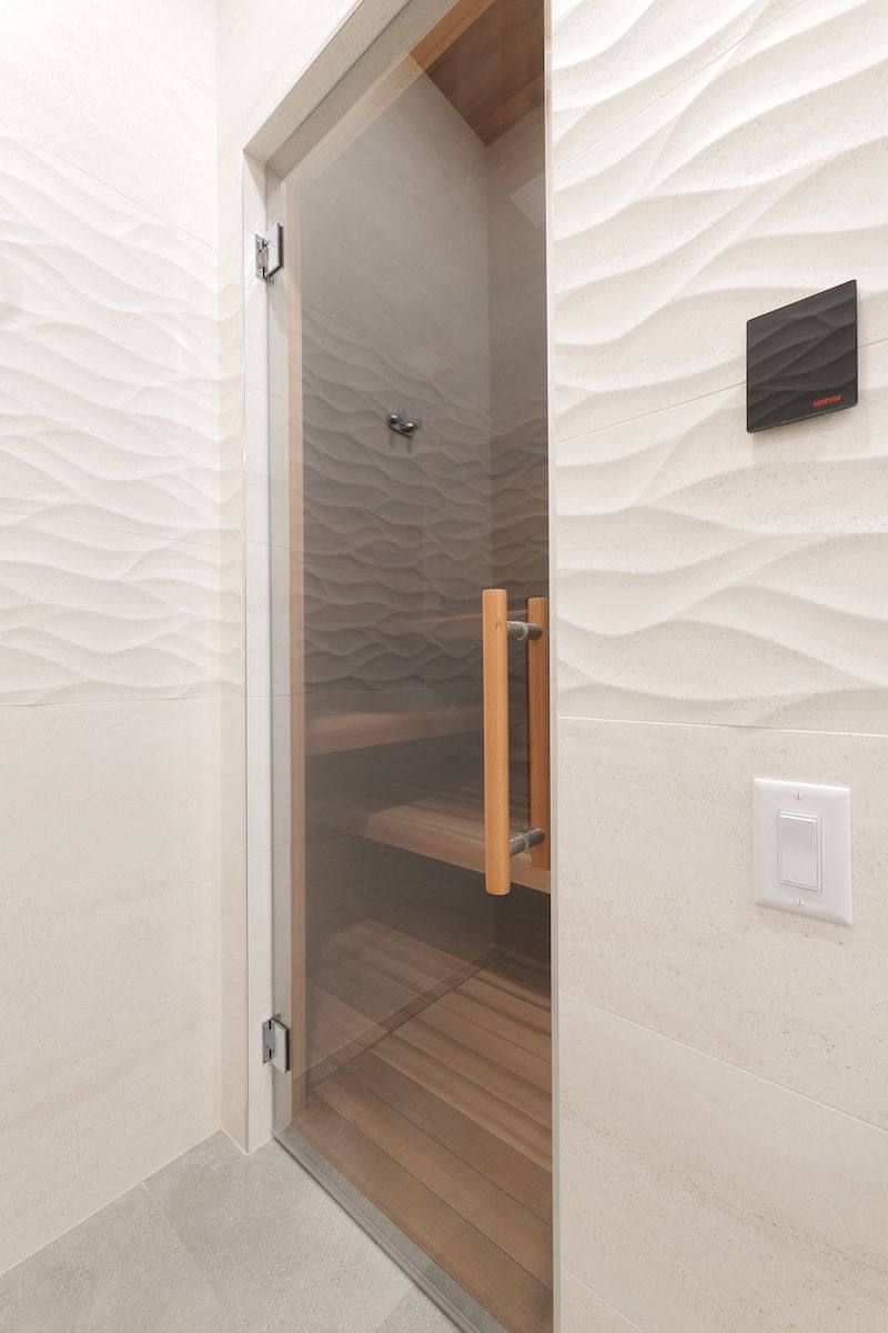 Glass door of a sauna with wooden interior and wavy white wall tiles.