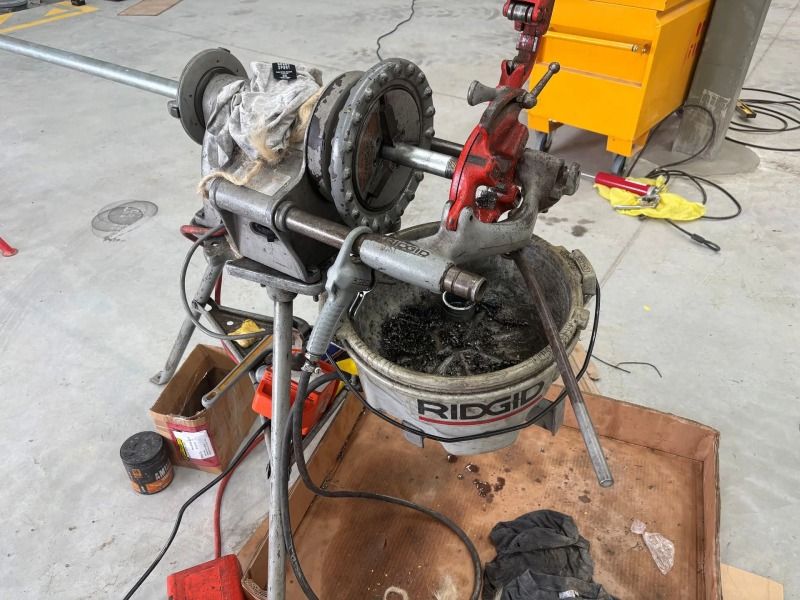 Pipe threading machine with metal pipes in a workshop, beside a white RIDGID bucket and tools on the floor
