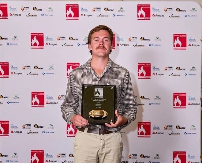 Man holding an award plaque and trophy at a step-and-repeat press backdrop.