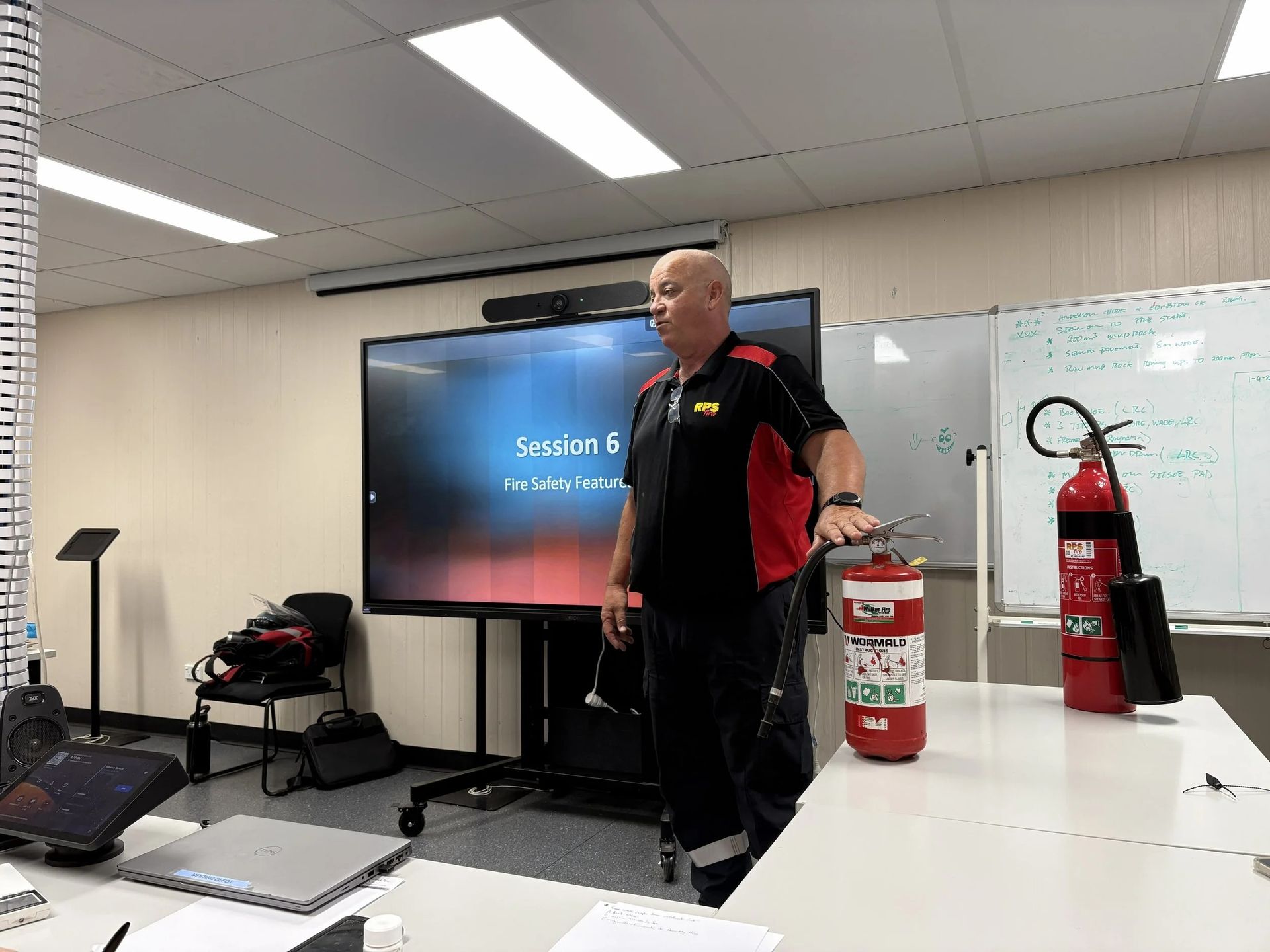 Presenter standing beside a TV screen in a conference room with a fire extinguisher in front.