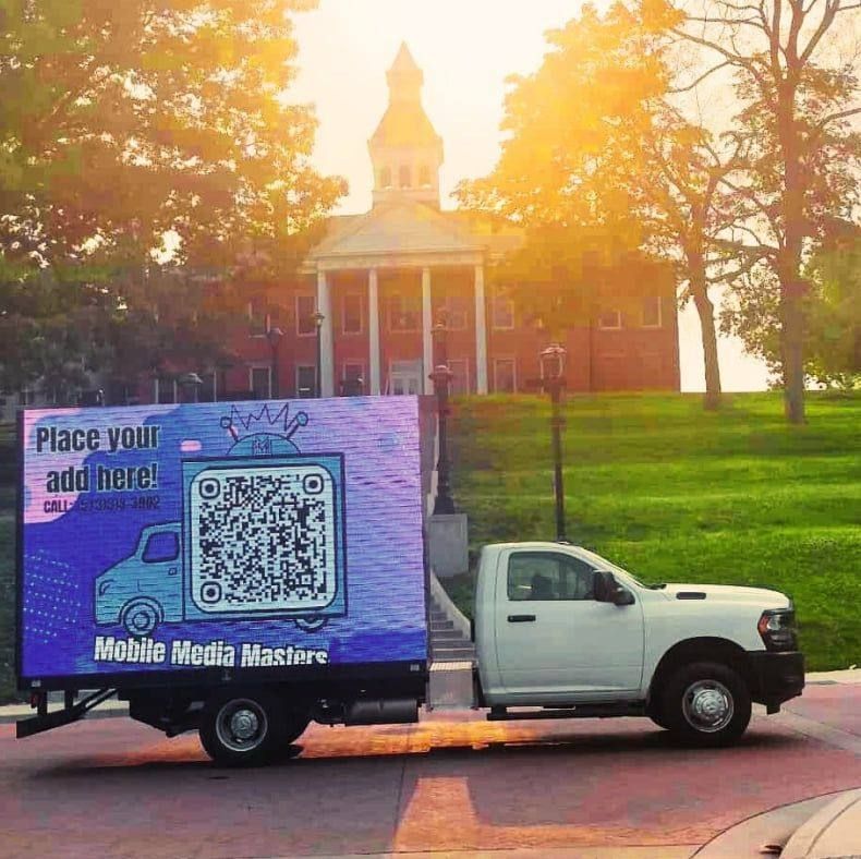 A digital billboard truck parked in front of a campus building, displaying a QR code and the text "Place your ad here!"