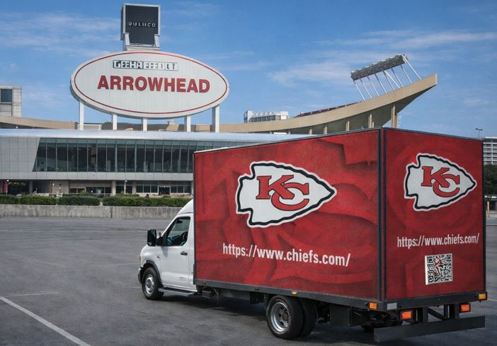 A white truck with red Chiefs branding parked in front of the Arrowhead Stadium sign under a blue sky.