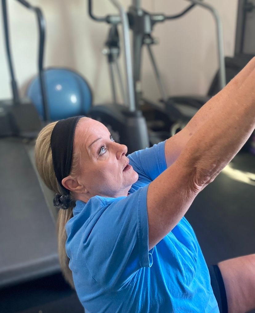 A woman in a blue shirt is stretching her arms in a gym.