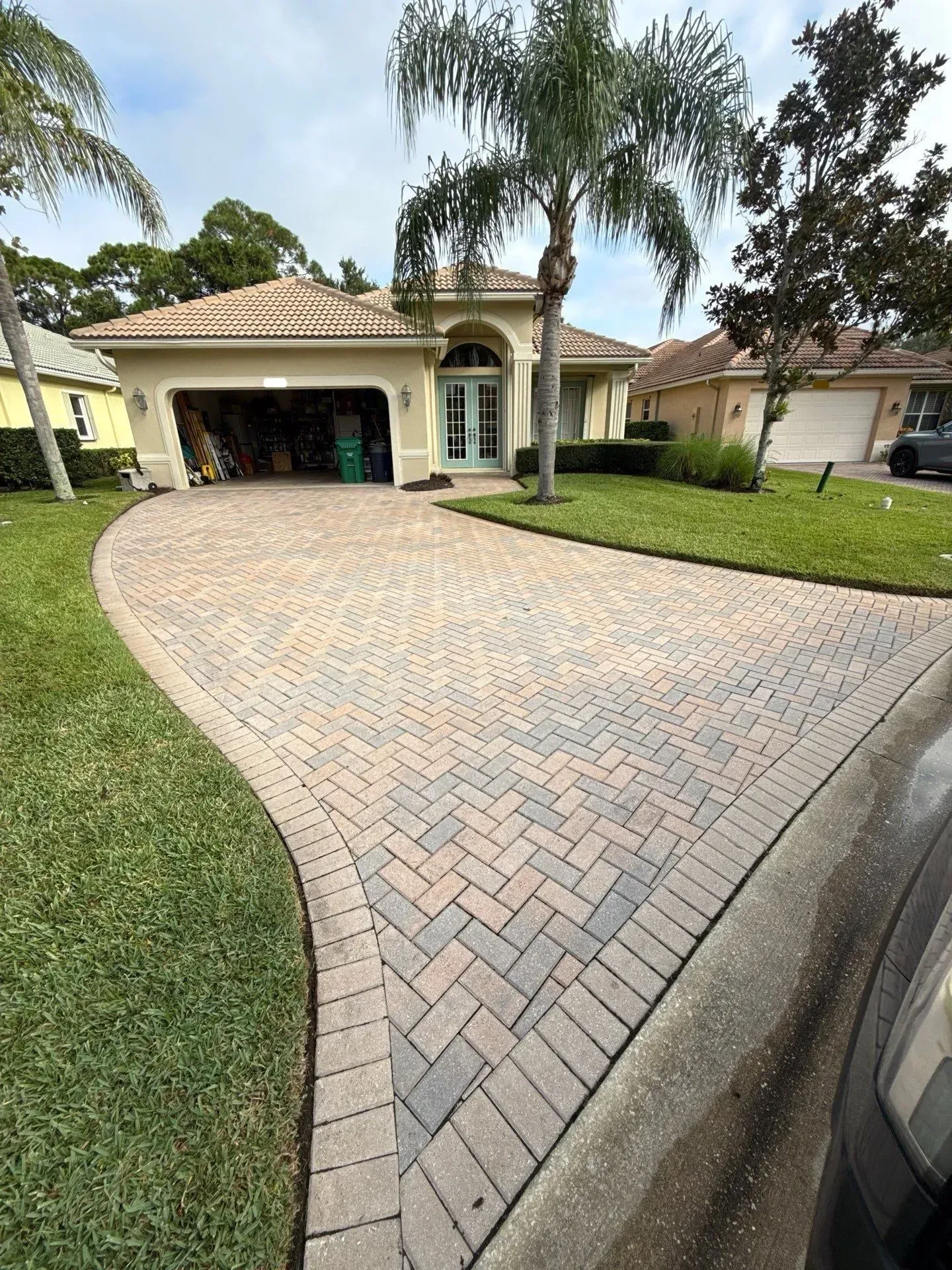 Curved brick driveway leading to a suburban house with a garage and palm trees in the front yard