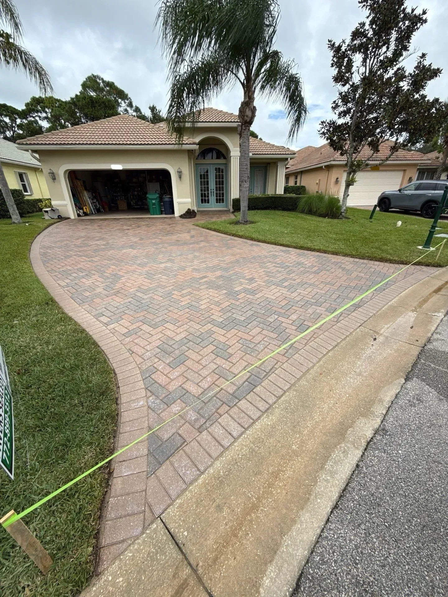 Brick-paved suburban driveway leading to a single-story house with garage and palm trees