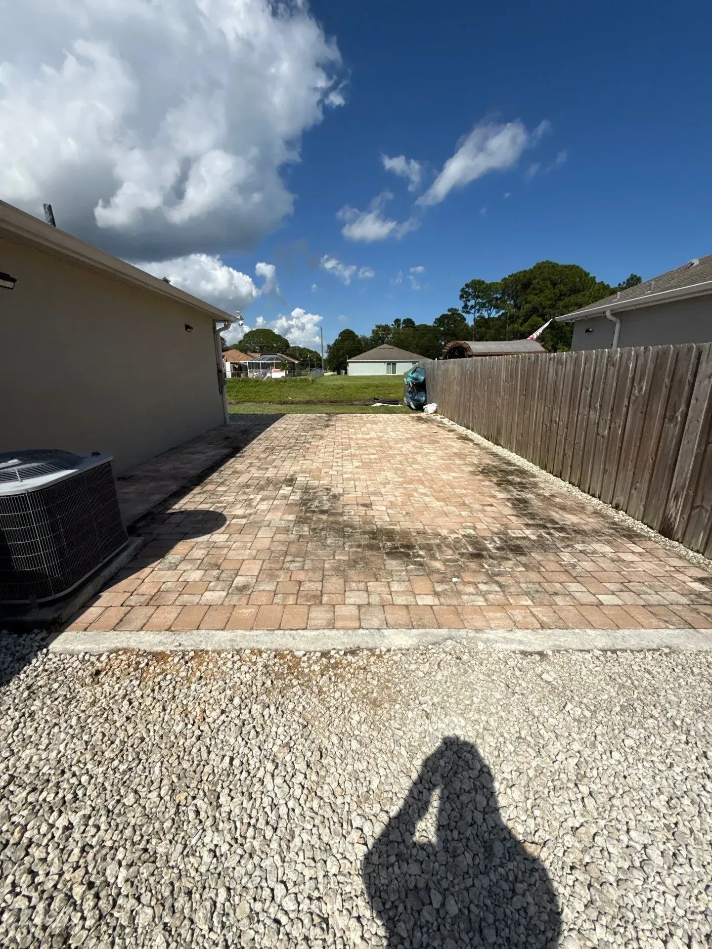 Backyard with brick patio and gravel path between a house and wooden fence under a blue sky
