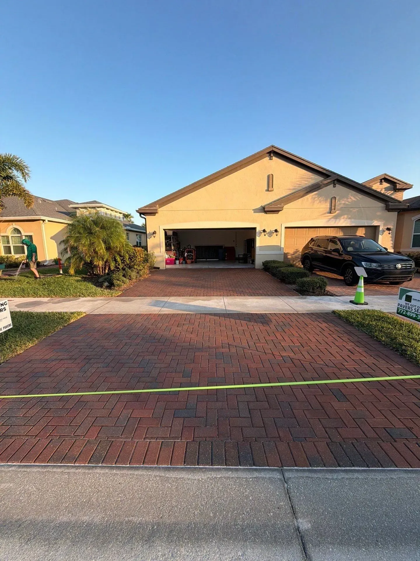 Suburban driveway with brick pavers, a tan house, parked cars, and lawn in the front yard.