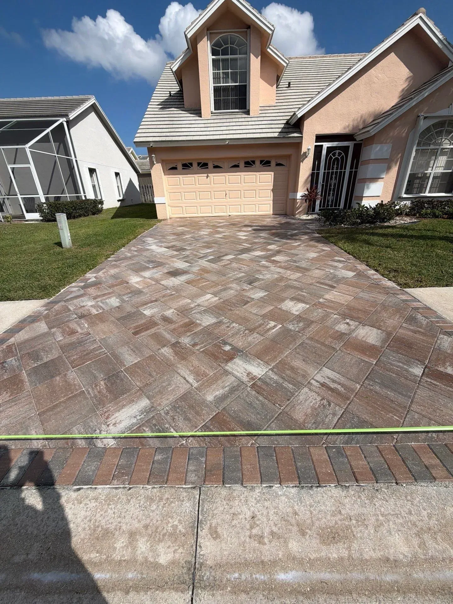 Brick driveway leading to a tan suburban house with a double garage and dormer window under a blue sky