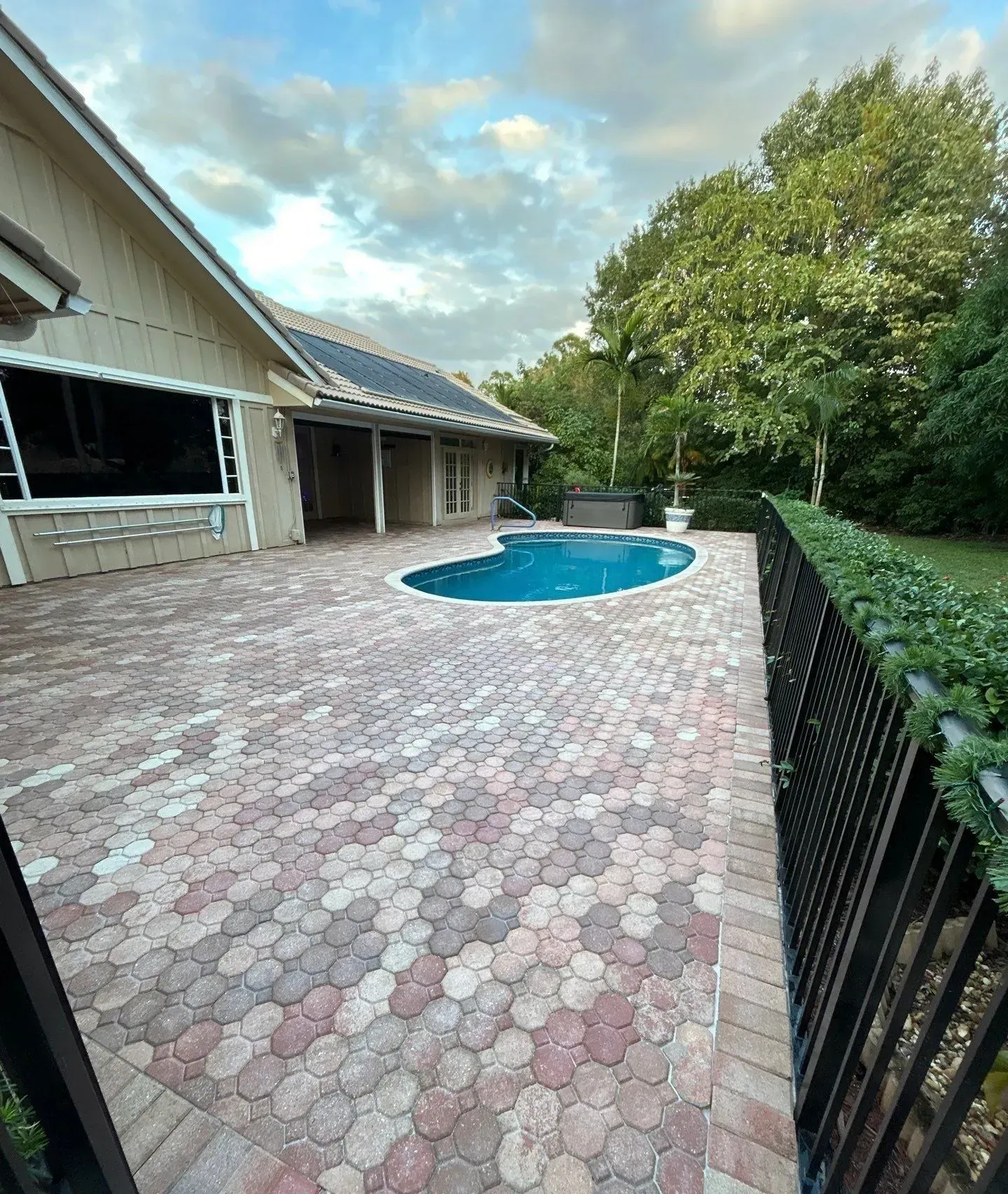 Backyard patio with a curved blue pool beside a house and trees, paved in pink and gray stone