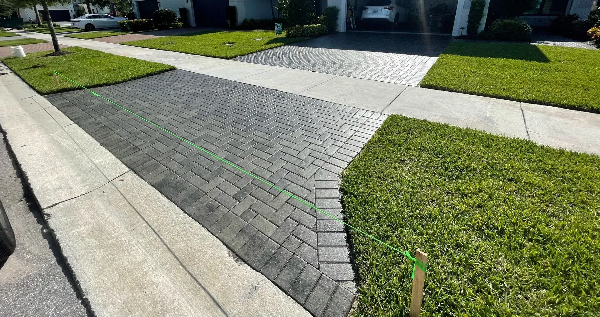Brick driveway with a pale concrete path crossing to a house, bordered by green grass and a curb.