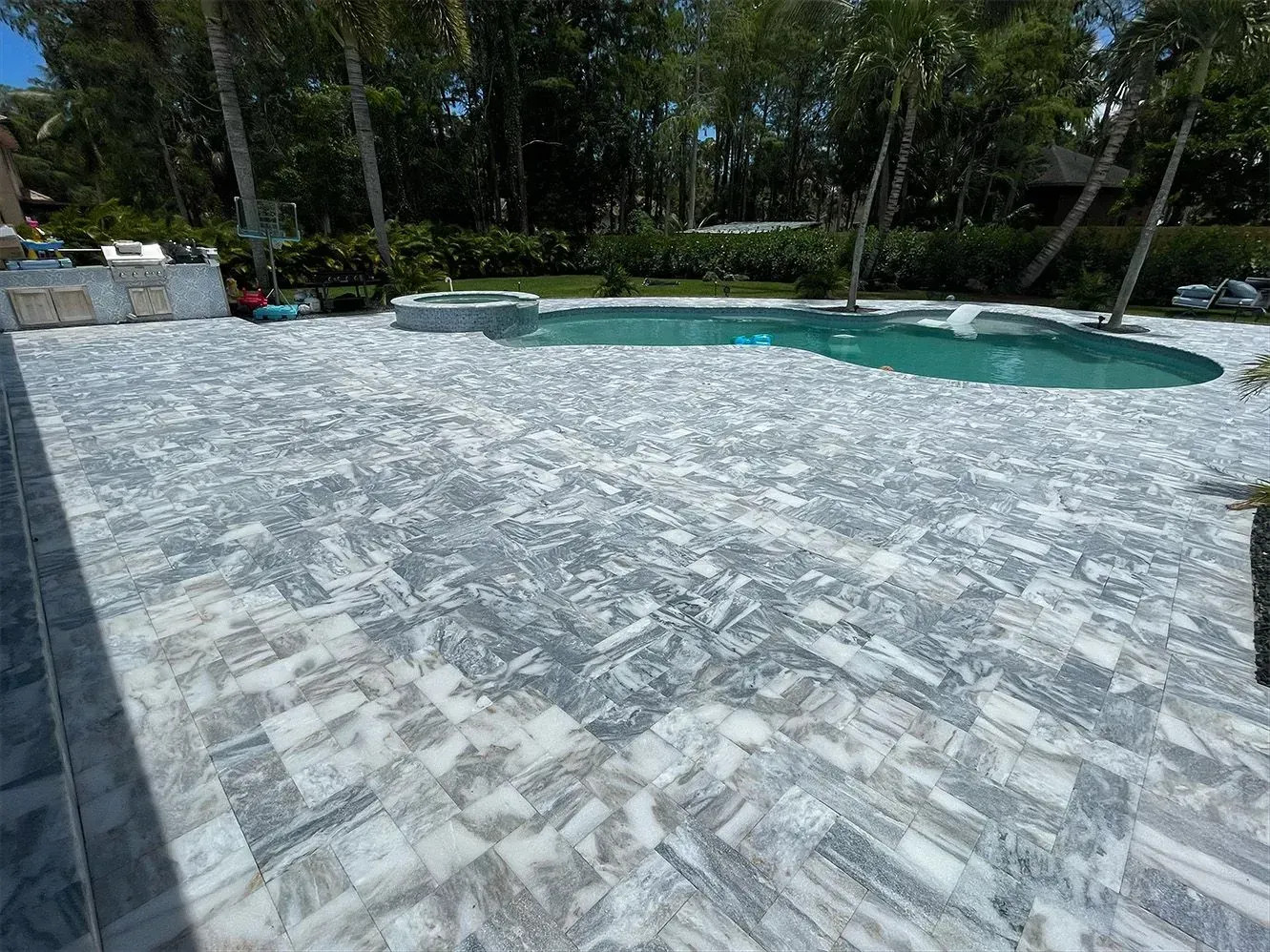 Stone-paved pool deck beside a turquoise swimming pool, surrounded by trees and greenery.