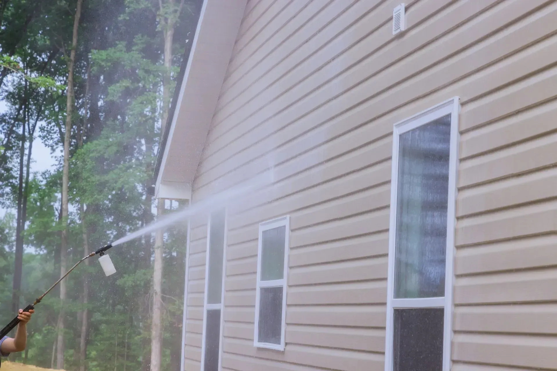 Person pressure-washing beige house siding outdoors with a long wand amidst mist and trees