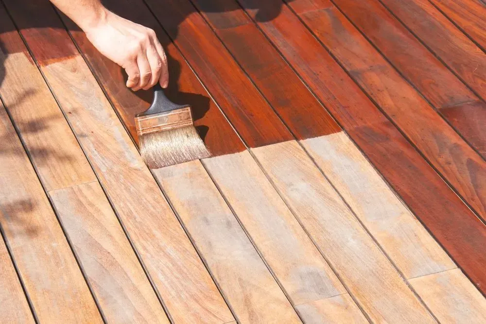 A hand paints a wooden deck, with one section already coated in light stain.