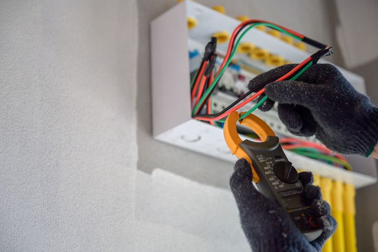 Gloved hands wiring an open electrical box with a multimeter in a wall cavity