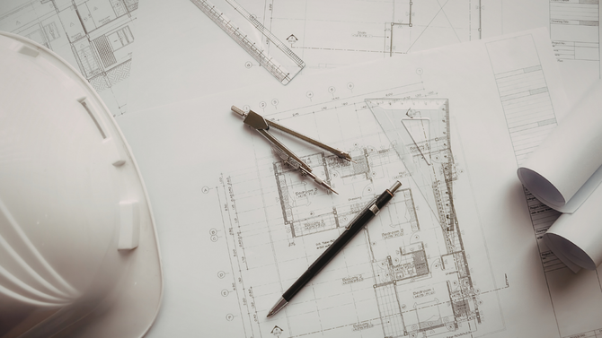 A white hard hat sits beside a technical drafting compass, a pencil, and rolled blueprints on a table.