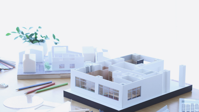 Architectural models of houses sit on a desk with pencils and a plant in a bright, minimalist workspace.