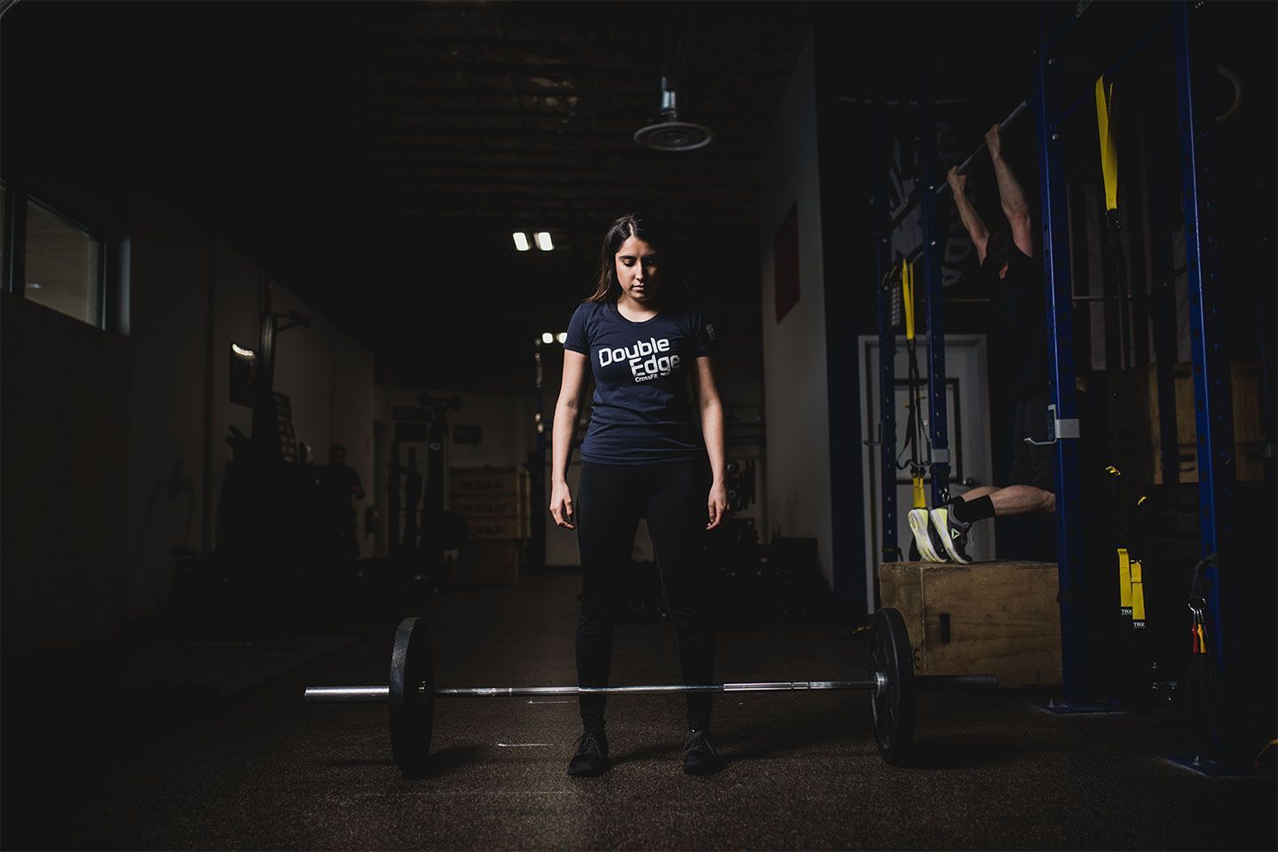 Person standing in a dim gym beside a barbell, wearing a dark shirt and pants.