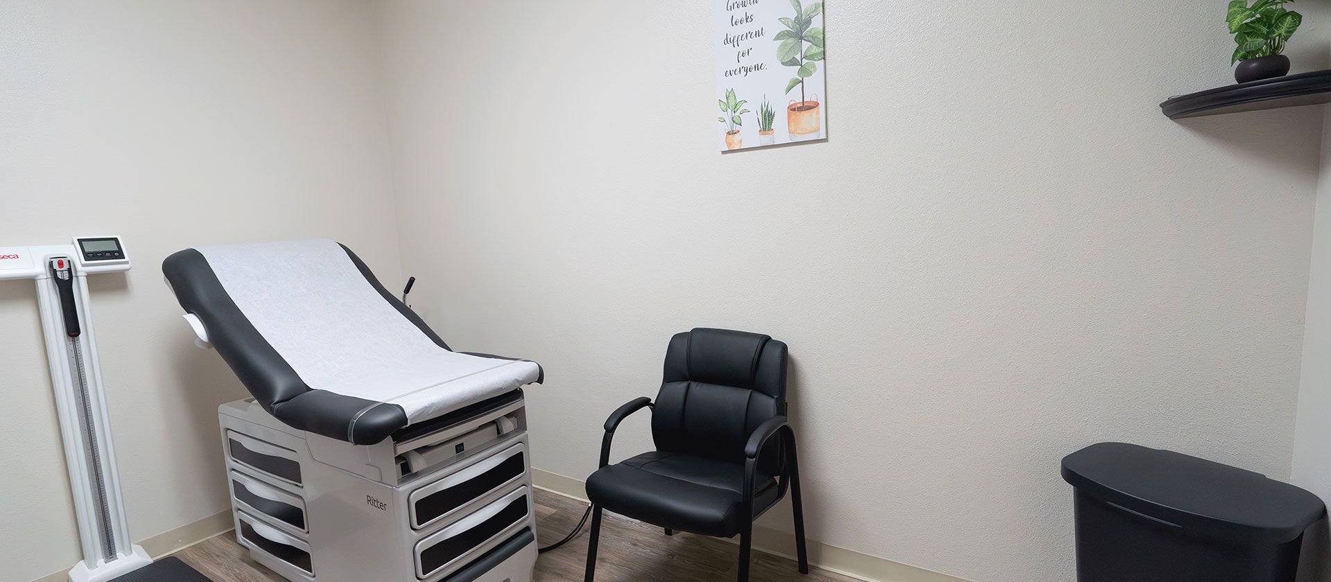 Medical exam room with exam table, chair, and stool against beige walls.