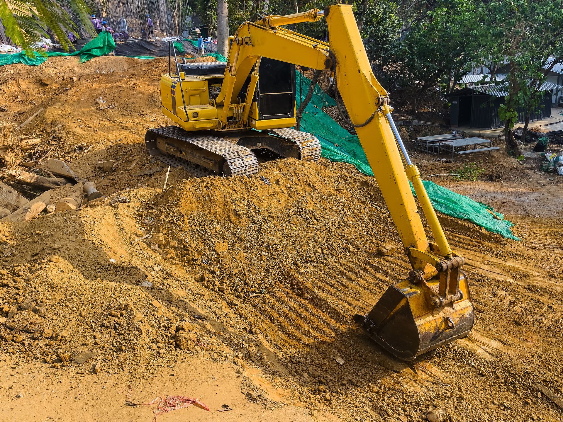 A yellow excavator performs land clearing near trees and a fence on a sloped construction site.