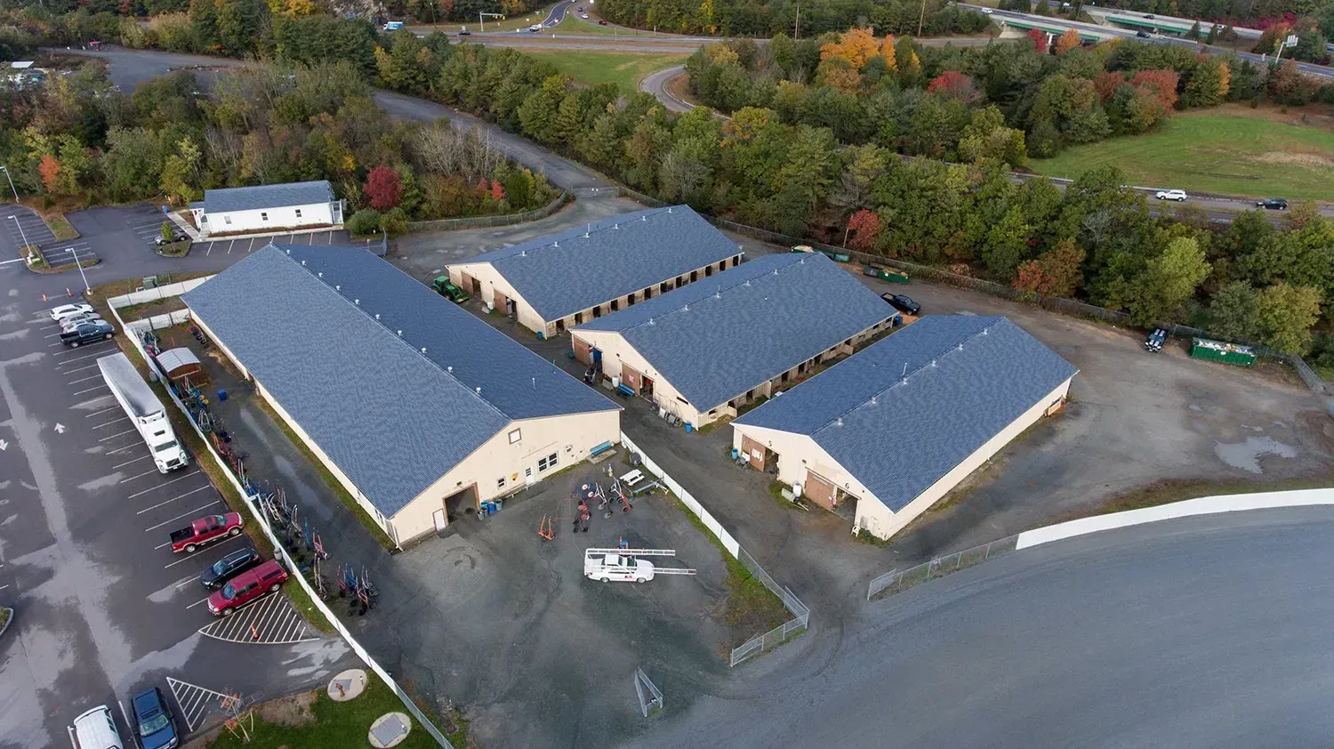 Aerial view of a large industrial building complex with blue roofs beside a curved road and trees.