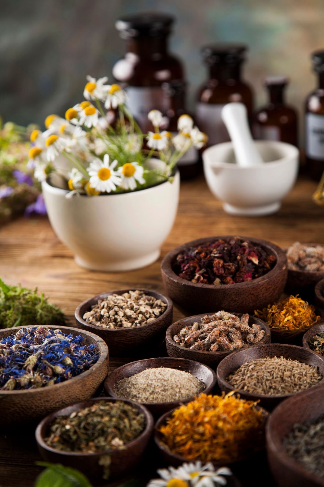 Un mortaio pieno di fiori di camomilla, circondato da ciotole di legno contenenti varie erbe essiccate e da boccette di vetro scuro da farmacia.